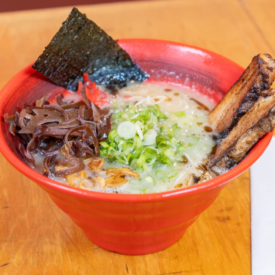 Red bowl of ramen topped with seaweed, wood ear mushrooms, green onions, garlic, and grilled pork ribs on a wooden table.