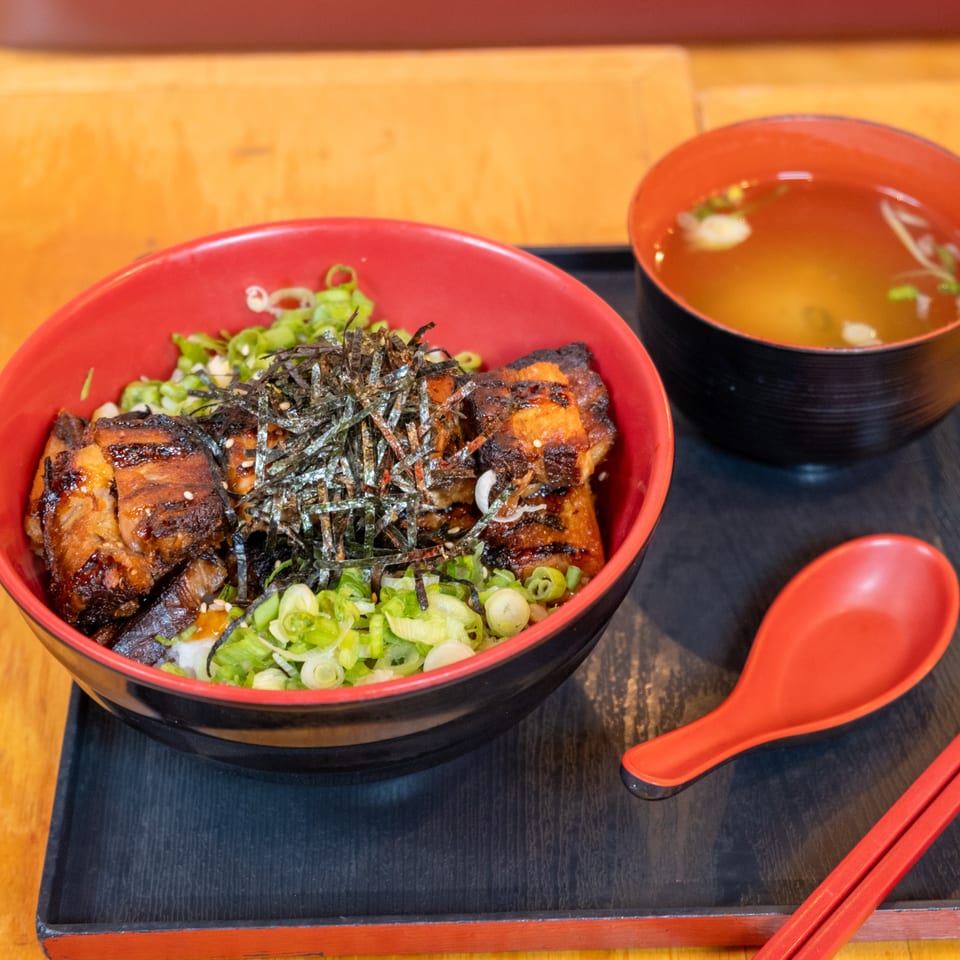 Japanese rice bowl topped with grilled meat, seaweed, and scallions, served with miso soup, spoon, and chopsticks.