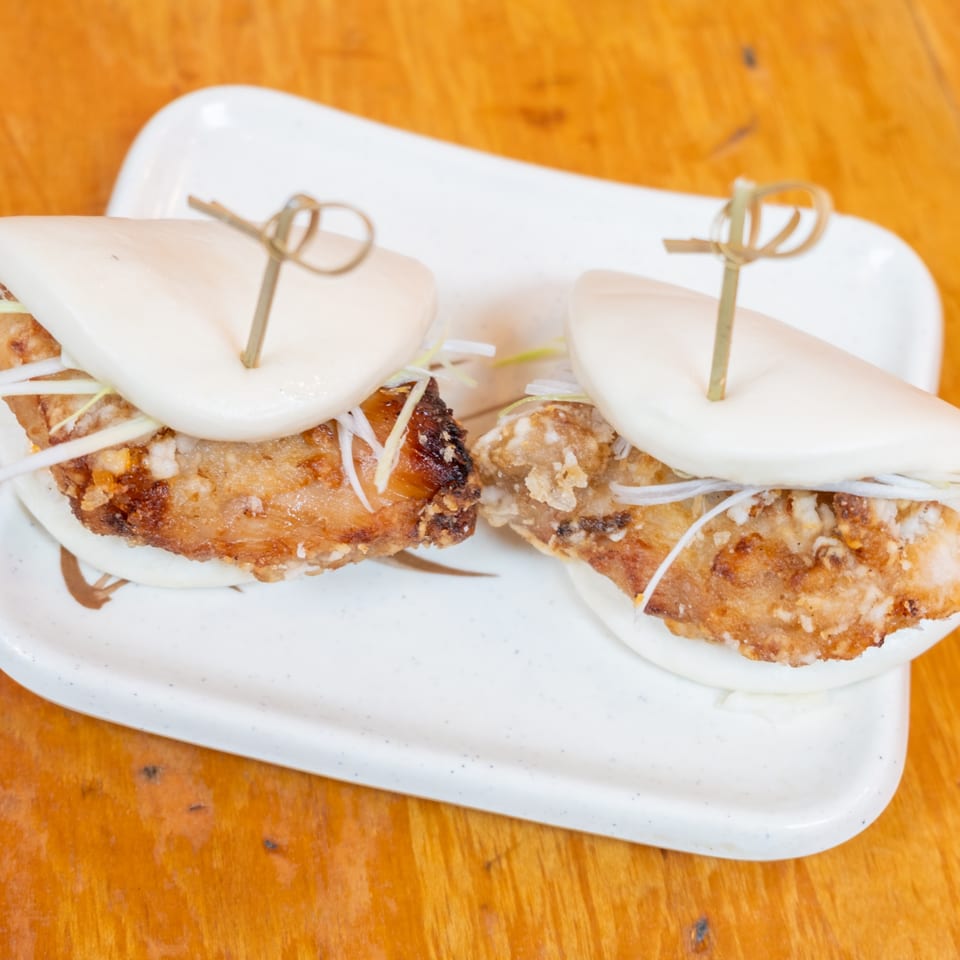 Two crispy fried chicken bao sandwiches with shredded vegetables on a white platter atop a wooden table.