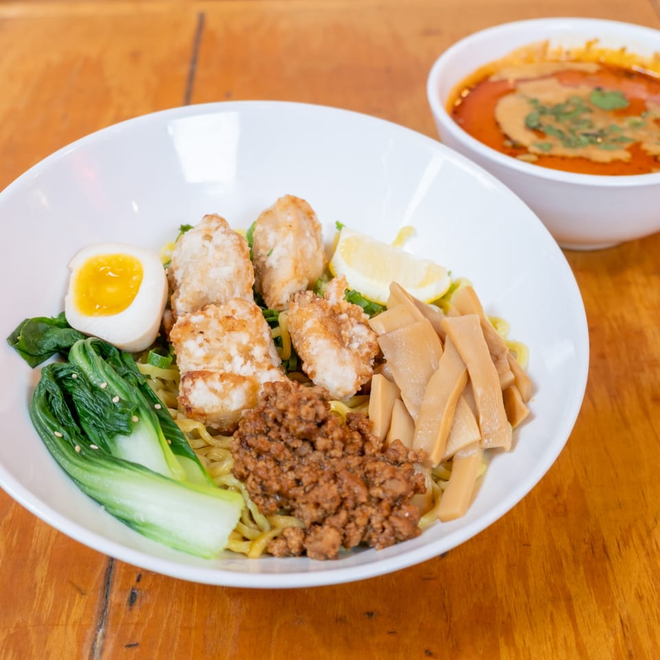 Bowl of noodles topped with fried chicken, minced meat, greens, bamboo shoots, and a soft-boiled egg, with spicy dip beside it.