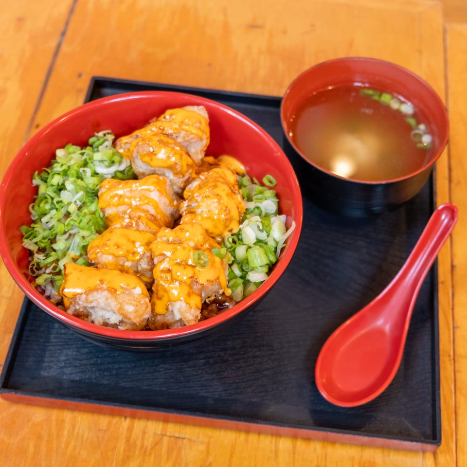 Japanese rice bowl topped with breaded chicken, orange sauce, and scallions, served with miso soup and red spoon on a black tray.
