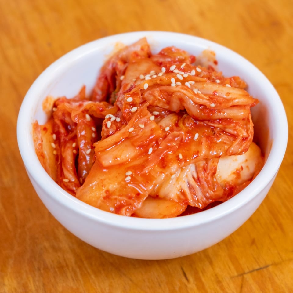 Spicy red kimchi topped with sesame seeds in a white bowl on a wooden table.